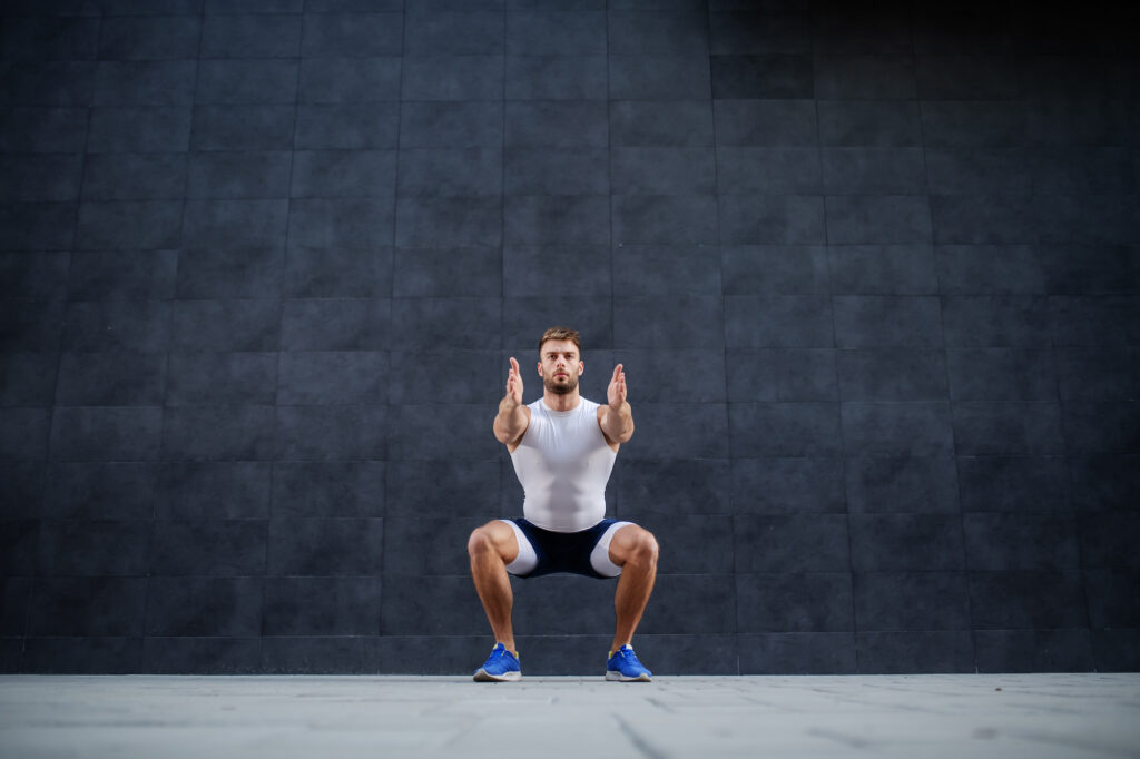 Handsome muscular caucasian man in shorts and t shirt doing squatting exercise outdoors. In background is gray wall.
