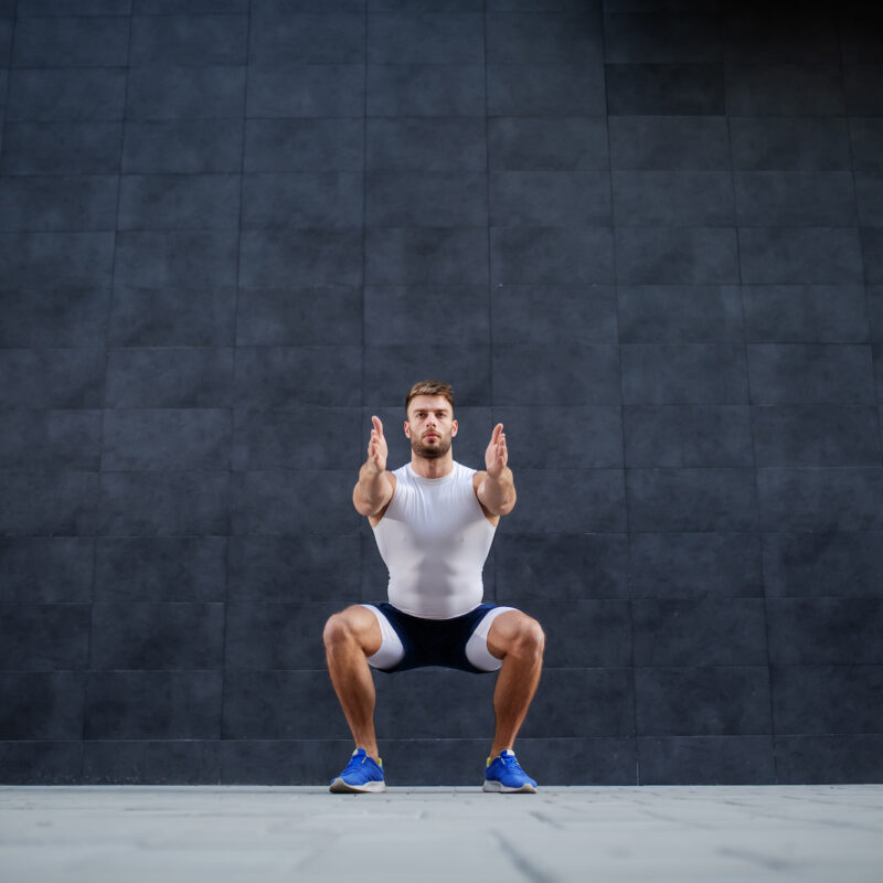Handsome Muscular Caucasian Man In Shorts And T Shirt Doing Squatting Exercise Outdoors. In Background Is Gray Wall.